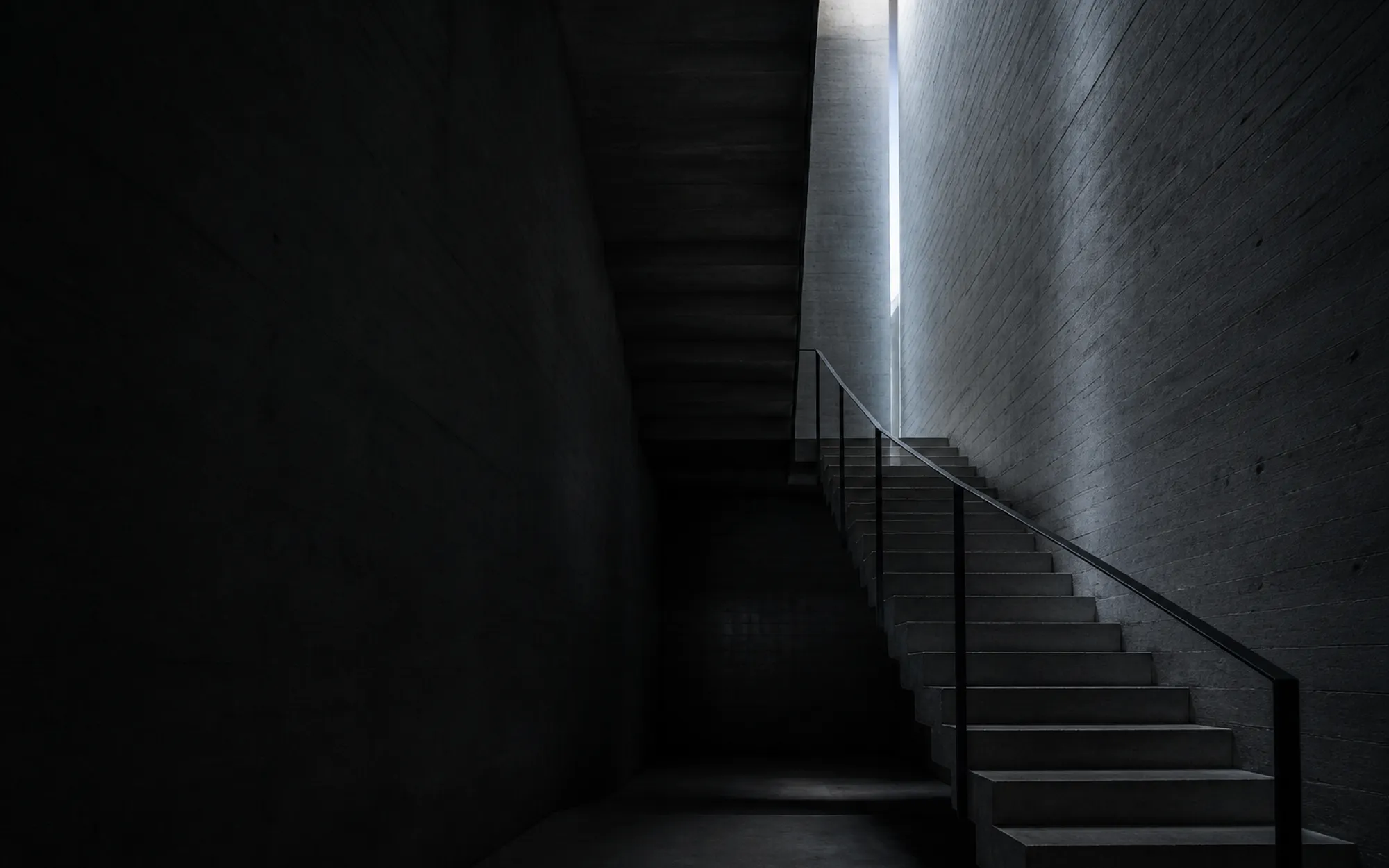 A cantilevered concrete stair turning through a tall board-marked void with a single slot of daylight