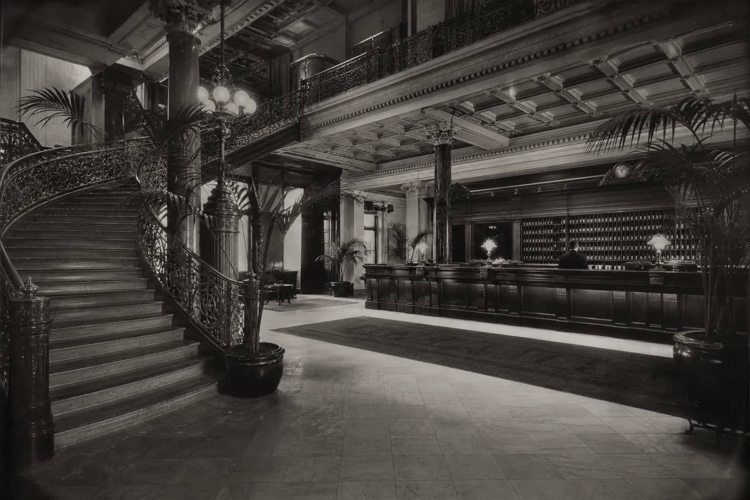 The lobby in an archival photograph: a sweeping cast-iron stair, potted palms, a long desk with brass rails and a wall of key pigeonholes behind it.